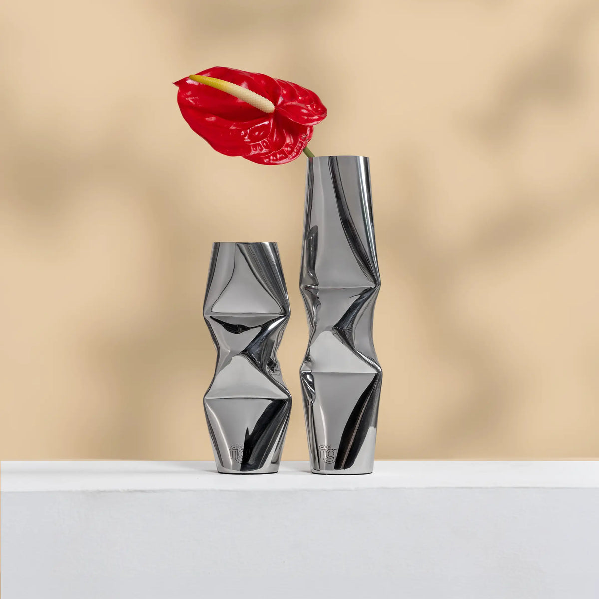 Two modern metallic silver chrome dented Vases with twisted geometric design, displayed on a white surface against a warm beige background. A red flower is placed in the taller vase.