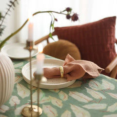 A styled dining table setting with a pale green and cream botanical tablecloth, a beige ceramic plate, a folded blush pink napkin, a gold ring, and two lit white candles in brass holders. A rust-colored chair and a textured burgundy pillow are softly blurred in the background.