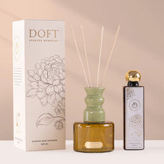 A reed diffuser set with a white floral Doft box, an amber glass diffuser bottle with a pale green neck and reeds, and a small dark refill bottle with a gold cap, arranged on a beige background.