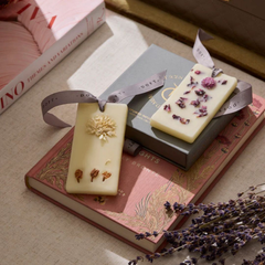 Two rectangular cream-colored Flower Blossom Wax Tablets with dried botanical elements and ribbons, styled on vintage pink books with lavender sprigs.