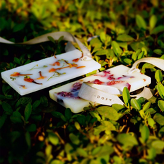 Two white rectangular Flower Blossom Wax Tablets with dried botanical inclusions and white ribbon loops, resting on green foliage. The brand name 'DOFT' printed on the ribbon.
