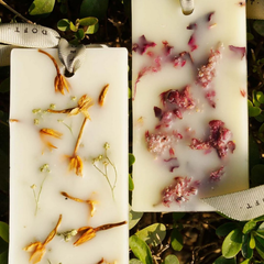 Two rectangular white resin Flower Blossom Wax Tablets with ribbon loops, each containing pressed dried flowers, shown side by side outdoors among green leaves.