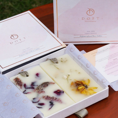 Two rectangular bars of DOFT natural wax candles in a white gift box, displayed on a wooden table with the matching pink and white packaging boxes behind them.