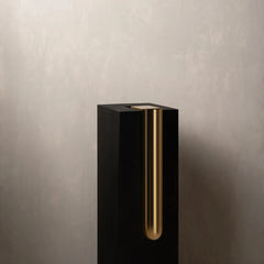 A modern black Bookcase Vase with a vertical brushed gold metal accent, set against a textured light gray wall.