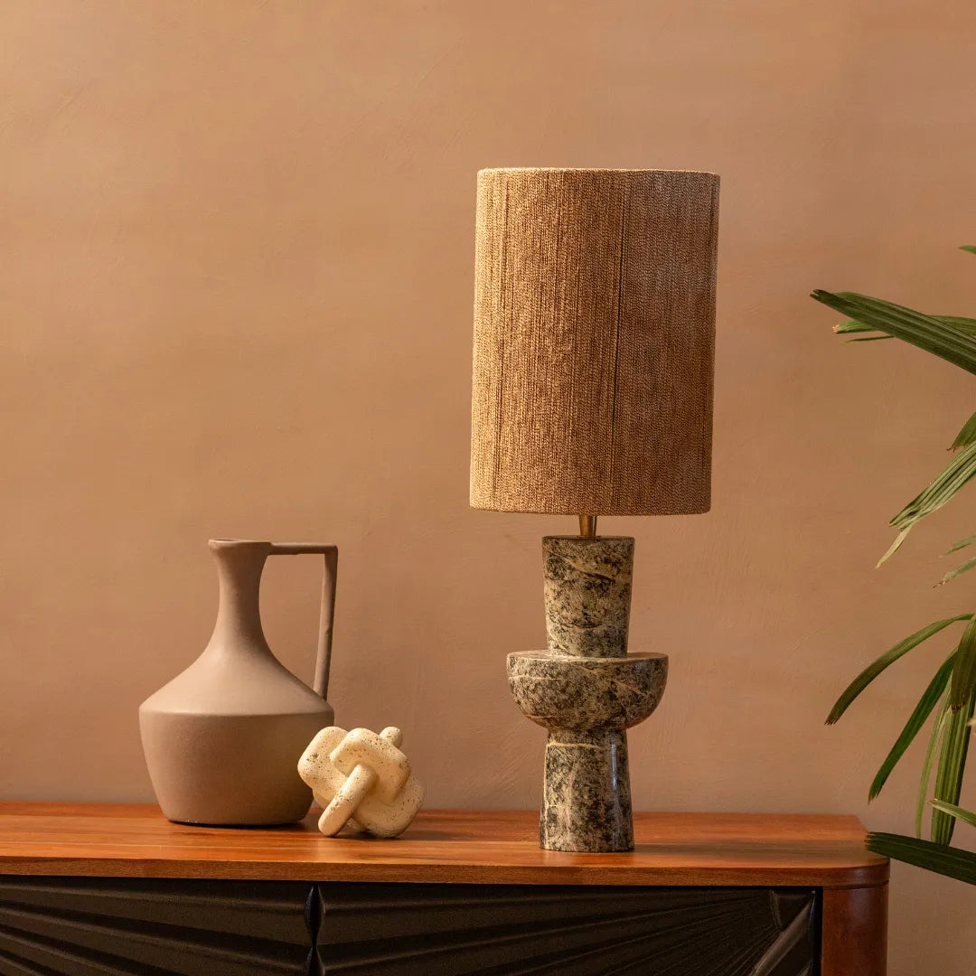 Natura Table Lamp with a textured shade on a wooden surface next to a vase and small sculpture