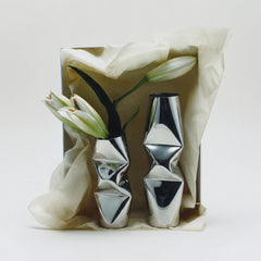 Two decorative metallic silver Chrome Dented Vases with sculptural, crumpled-folding designs, displayed in a gift box lined with cream tissue paper and decorated with white lilies, on a light gray background.