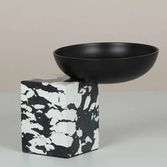 A matte black ceramic bowl resting on a black and white marbled stone pedestal.