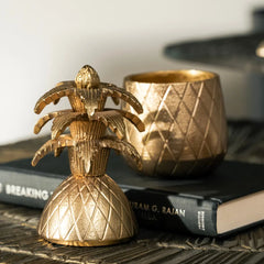 Decorative Gold pineapple-shaped candle votive on a surface with books and another gold item in the background.