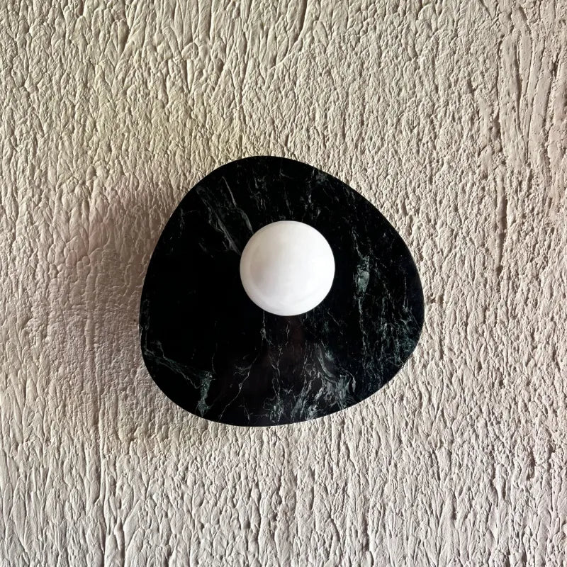 Modern wall-mounted light fixture featuring an organic, irregularly shaped black green marble base with a central white spherical element, mounted on a textured off-white plaster wall.