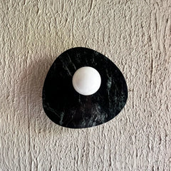 Modern wall-mounted light fixture featuring an organic, irregularly shaped black green marble base with a central white spherical element, mounted on a textured off-white plaster wall.