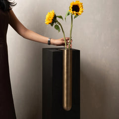 A person holding two yellow sunflowers in a tall, cylindrical brushed brass Bookcase Vase