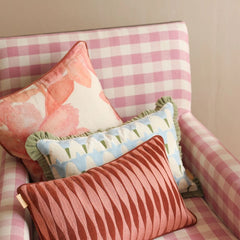 Three decorative throw pillows arranged on a pink and white gingham armchair. The pillows include a large pink floral print pillow, a light blue and white patterned pillow with green ruffles, and a rust pink pillow with a wavy striped texture.