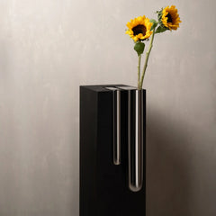 A modern black rectangular pedestal vase with two vertical chrome tubes, holding two yellow sunflowers.