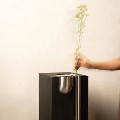 A hand holding a long stem of pale yellow flowers inserts it into a modern black rectangular vase with a brushed silver metallic bowl-shaped opening.