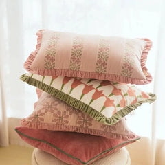 A stack of four decorative throw pillows with ruffled edges, featuring floral and geometric patterns in soft pink, peach, green, and cream tones, set against a bright, softly lit background.