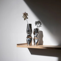 Two metallic sculptural Chrome Dented Vases with a twisted, hourglass-like geometric design, displayed on a light wood floating shelf.