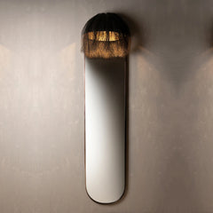 A modern wall-mounted mirror with a long, narrow oval shape, featuring a black fringed lampshade-style top and a warm light glowing from within.