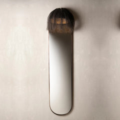 A modern wall-mounted mirror with a long, narrow oval shape and a dark, shaggy textured top section.