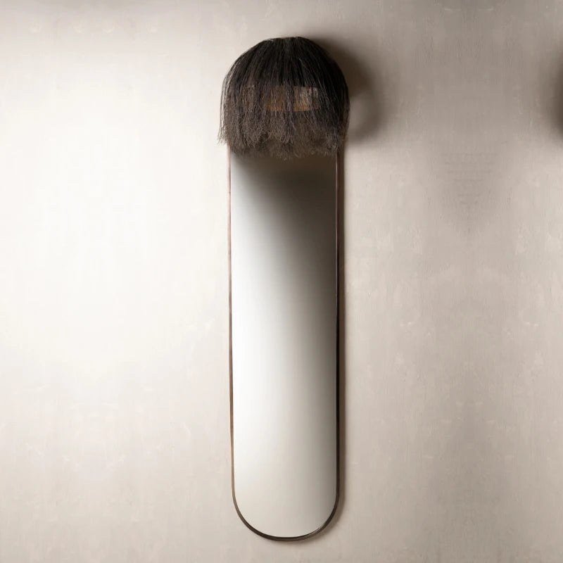 A long wall mirror with a long, narrow oval shape and a dark, textured, shaggy top section.