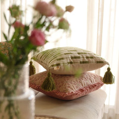 Two decorative throw pillows stacked on a light upholstered bench in a softly lit room. The top pillow is cream with olive green stripes and tassels, and the bottom pillow is dusty pink with a textured pattern and ruffled edge.