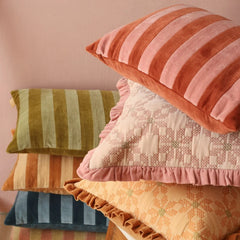 A stack of decorative throw pillows in soft, coordinated fabrics and colors, including rust pink, olive green, cream, peach, and navy blue, set against a dusty pink wall.