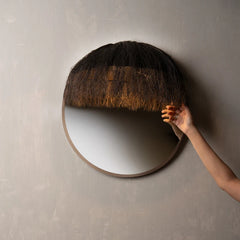 A round wall mirror with a dark brown textured hair-like top and a light wood frame.