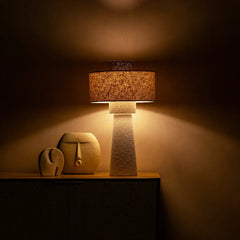 Iris Table Lamp with textured base and wicker shade casting a warm glow on a dark surface.