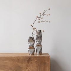 Two modern Chrome Dented Vases with dried floral branches, displayed on a light brown wooden sideboard against a neutral off-white wall.