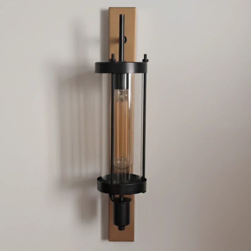 A modern industrial Edison Column wall sconce with a black metal frame, clear glass cylinder, and warm Edison bulb, mounted on a vertical rectangular brass plate against a neutral beige wall.