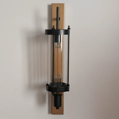 A modern industrial Edison Column wall sconce with a black metal frame, clear glass cylinder, and warm Edison bulb, mounted on a vertical rectangular brass plate against a neutral beige wall.