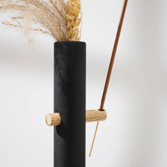 Black vase with wooden accents holding dried plants and incense holder on a white background