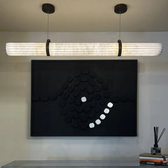 A modern horizontal linear pendant light with a ribbed, translucent white shade and black hanging hardware, suspended above a black wall art piece in a styled interior setting.