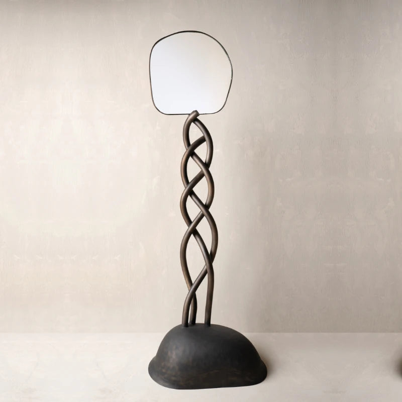 A sculptural floor mirror with a dark twisted metal stand and a rounded base.