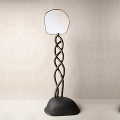 A sculptural floor mirror with a dark twisted metal stand and a rounded base.