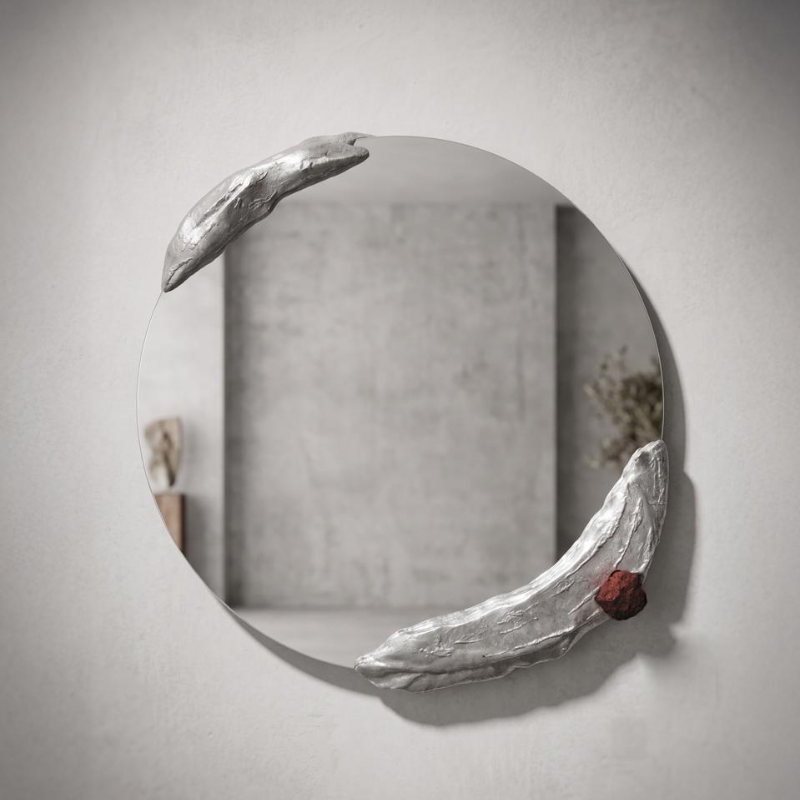 A round wall mirror with a sculptural, organic silver frame resembling two curled leaves or branches, accented with a small textured red piece, mounted on a light gray wall.
