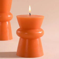 An orange Pillar candle (Non Scented) with a lit flame, set against a warm beige background with a second candle blurred in the background.