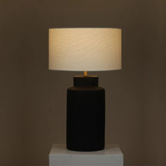 Jasper Table Lamp with a black base and beige lamp shade on a white pedestal against a dark background