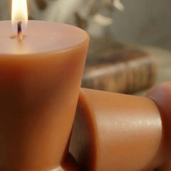 A close-up of a lit wax surface in peach-colored pillar candle with another matching candle resting beside it on a dark surface.