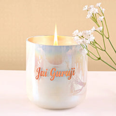 A lit white iridescent U-Glass candle jar with Flower Blossom Scent and the words 'Jai Guruji' in orange script on the front, set on a light surface against a beige background.