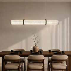 Linear pendant light with a ribbed glass shade and dark metal end caps, suspended above a wooden dining table.