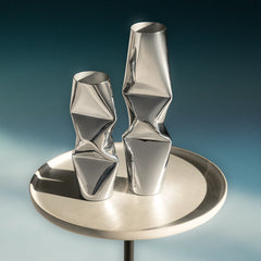 Two sculptural metallic vases with dented design in chrome, twisted geometric shapes displayed on a round brushed-metal pedestal against a blue gradient background.