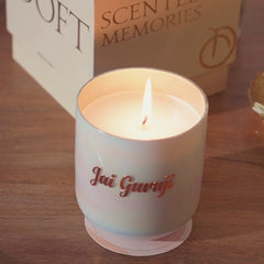 A lit scented U-glass candle in a frosted white glass jar with the brand name 'Jai Guruji' printed in rose gold script, sitting on a wooden surface with its branded packaging box. 