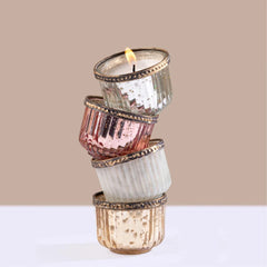 Gilded Votive -Four stacked glass candle holders in silver, rose gold, ivory, and blush with gold trim, with the top candle lit, set against a beige background. 