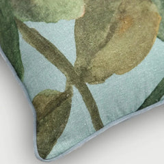 Close-up of a decorative throw pillow corner showing a botanical print with muted green and olive leaves on a pale blue background, finished with a light gray stitched edge.