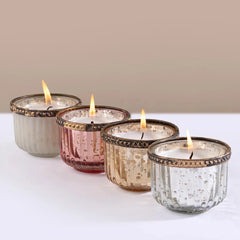 Gilded Votive Four lit candle holders in ribbed glass with metallic finishes, arranged diagonally on a white surface against a beige background. 