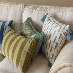 Three decorative throw pillows arranged on a cream-colored sofa. The front-left pillow has mustard yellow and cream vertical stripes, the front-right pillow has blue and cream vertical arrow-like stripes with a blue tassel, and the back pillow has a green botanical pattern with a light blue border and a blue tassel.