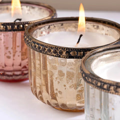 Three lit glass Gilded Votive Candle set, with fluted ribbed sides and metallic trim, shown close up on a light surface. The center holder is champagne gold with dark metallic detailing, and the side holders are pink and clear.