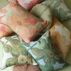 A styled collection of floral throw pillows in various colors and patterns, including green, peach, pink, and light blue, arranged together on a pale green surface.