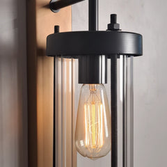 Industrial-style wall sconce with a black metal frame, clear glass cylindrical tubes, and a glowing filament light bulb.