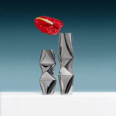 Two modern metallic silver vases with a faceted, sculptural design in chrome, one short and wide, the other tall and narrow, displayed on a white surface with a red anthurium flower in the taller vase.